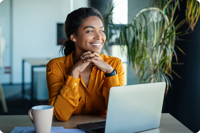 Portrait of dreamy african american businesswoman sitting at desk in office and thinking while working on laptop, looking away with pleased face expression,